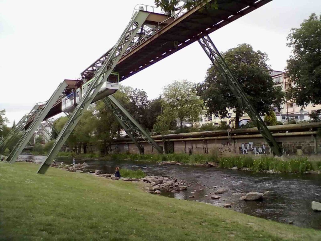 Wuppertaler Schwebebahn: Blick von unten vom Ufer aus auf die Schwebebahn Konstruktion mit einer anfahrenden Bahn.